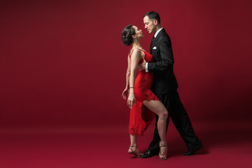 Couple of professional tango dancers in elegant suit and dress pose in a dancing movement on red background. Handsome man and woman dance looking  eye to eye. - obrazy, fototapety, plakaty