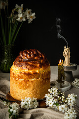 Homemade Ukrainian traditional Easter bread -- paska, natural bee wax candles and white spring flowers on linen tablecloth on dark background.