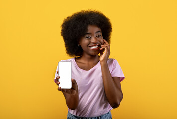 Mobile app advertising. Pretty black bushy woman showing smartphone with blank screen on yellow studio background