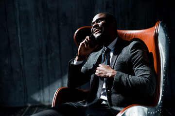 Stylish african american businessman sits on a chair in the office and communicates on the phone smiling - Powered by Adobe