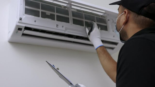 Air conditioner service indoors. Asian technician checking list and fix the Air conditioner. Air conditioner cleaning technician Clear the blocked air filter. He in uniform wearing rubber and mask.