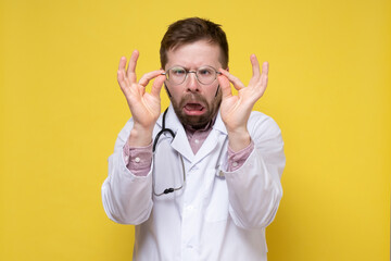 Perplexed doctor with poor eyesight has taken off glasses and is examining them with a stupid expression on face. Yellow background.