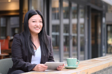 portrait of smiling Asian young business woman using digital tablet at outdoor cafe, looking at camera