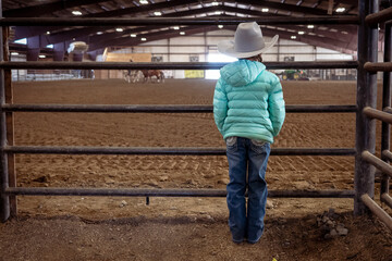 A little girl with a big cowboy hat, light blue down and sparkling rodeo jeans watches through the iron gate of an rodeo arena as riders practice for rodeo. - obrazy, fototapety, plakaty