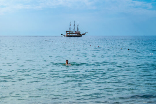 A man floats in the clear blue waters of the Mediterranean Sea and a vintage three-masted pirate ship on the horizon. Seascape on the beach in Alanya (Turkey). Background with copy space - Powered by Adobe