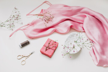 Composition with pink silk fabric, gift in craft paper and threads and scissors on white background. Flat lay, top view. Hobby, leisure concept.