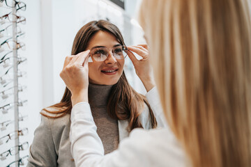 Beautiful and fashionable woman choosing eyeglasses frame in modern optical store. Female seller...