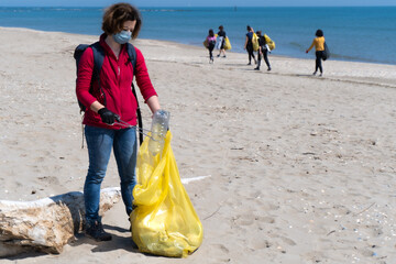Donna che pulisce la spiaggia mettendo una bottiglia di plastica nel sacco dei rifiuti, a Lido di Dante in Italia, e sullo sfondo ci sono altre persone che portano sacchi di rifiuti. Senza plastica.  - obrazy, fototapety, plakaty