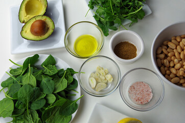 Healthy food ingredients used to prepare a delicious dip, including avocado, olive oil, garlic, cumin, pink salt, beans, spinach, cilantro and lemon