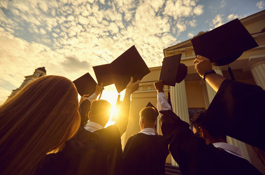 Low angle back view of company of graduated people in gowns standing with caps in raised hands near university at sunset and celebrating graduation