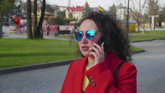 Beautiful young curly girl in a red coat and sunglasses chats on a smartphone in the city