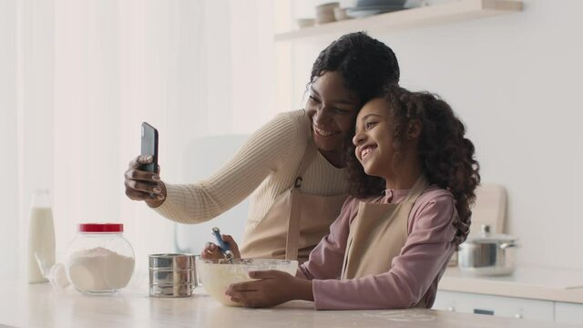 Happy african american mother and daughter making selfie at kitchem, smiling to smartphone while cooking bakery together