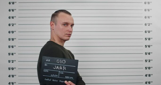 Mugshot of male serious criminal getting photographed at police station. Crop view of man in 30s turning head and looking to camera while standing aside and holding sign. Concept of crime.