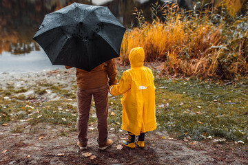 A little boy in a yellow raincoat with his father are walking in an autumn park with a black...