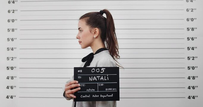Mugshot of young woman with ponytail turning to sides while holding sign and posing for photo . Crop view of female criminal looking to camera in police department. Concept of crime.