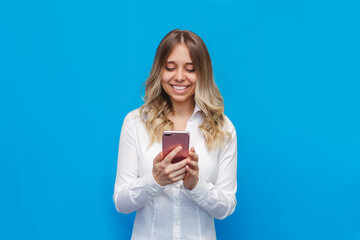 A young caucasian happy pretty blonde woman in a office white shirt smiles and holds a mobile phone looking at the screen isolated on a bright color blue background. Student girl uses her smartphone 
