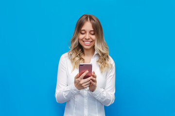 A young caucasian happy pretty blonde woman in a office white shirt smiles and holds a mobile phone...