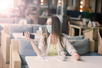 Serious woman with protective face mask looking at smart phone checking news on a cafe terrace