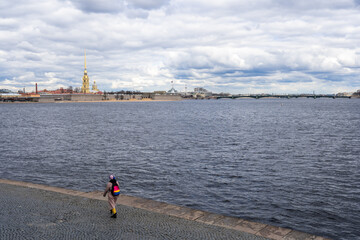 Russia, St. Petersburg, Vasilievsky Island. Granite embankment. Cloudy spring day. On the horizon is spire of the Peter and Paul Fortress against backdrop of grey clouds. Girl shooting by smartphone.