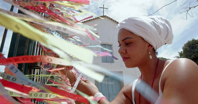 Brazilian woman dressed in the traditional Bahian costume of the Umbanda religion saying a prayer. Colored ribbons of religious promises in devotion to Senhor do Bonfim in Bahia, Brazil. 6K.