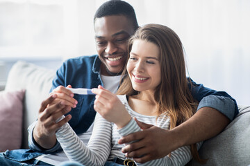 Happy young interracial couple holding pregnancy test, looking at positive result