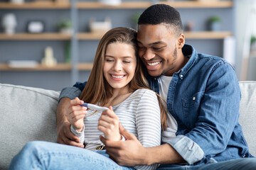 Happy multicultural couple holding positive pregnancy test and embracing on couch