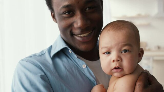 Close up portrait of happy african american daddy carrying and hugging his cute newborn baby, smiling to camera at home