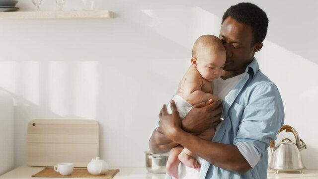 Young african american father carrying and calming his newborn baby wearing diaper, standing at kitchen, free space