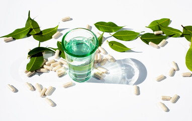 Glass cup with water and capsules and pills on a white background. Green leaves The topic of nutraceuticals and supplementation. Alternative Medicine Copy space for text.