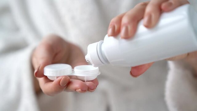 Closeup female hands are holding white plastic container for contact lenses. Woman is pouring cleaning liquid water for washing soft optical lens. Vision correction of myopia, hyperopia concept.