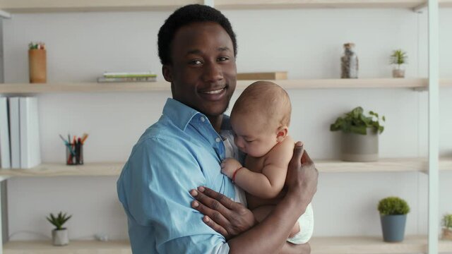 Close up portrait of happy african american dad carrying his newborn baby and smiling to camera, infant wearing diaper