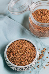 Organic uncooked scattered buckwheat grain in a bowl and glass jar on a light blue background