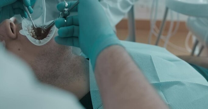 Close-up an Arabian patient with optragate in the mouth is having a dental treatment by using a dental mirror and injection while a female assistant is holding a saliva ejector.