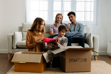 Cute little caucasian boy putting things like clothes in cardboard box for donation with help of teenage sister while happy mother and father watching them at home