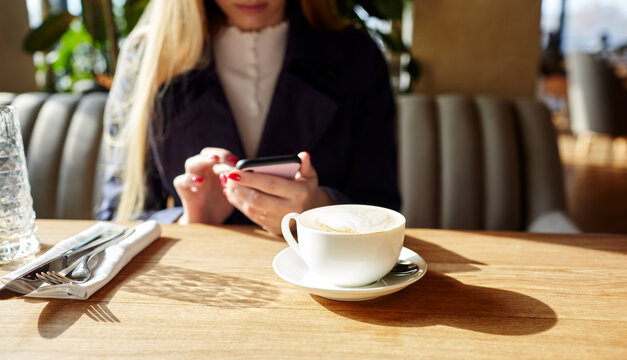 Woman typing text message on smart phone in a cafe. Young woman sitting at a table with a coffee using mobile phone. Coffee break. Cup of coffee on the table. Blurred image, selective focus
