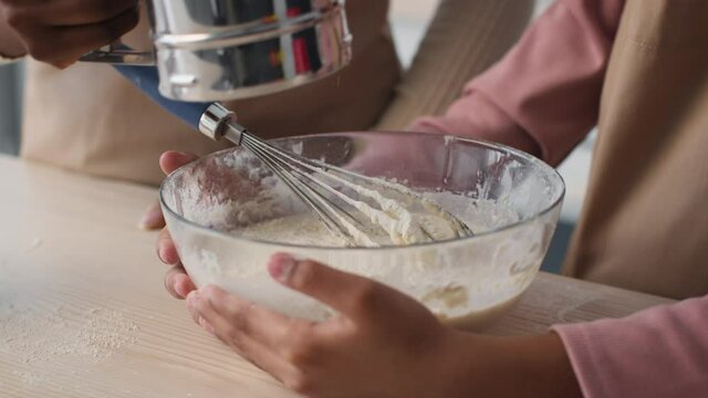 Close up of little black girl cooking pastry with mother, woman adding flour and daughter whisking dough at kitchen