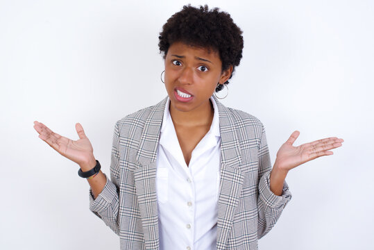 Clueless African American businesswoman with curly bushy hair wears  formal clothes over white background shrugs shoulders with hesitation, faces doubtful situation, spreads palms, Hard decision