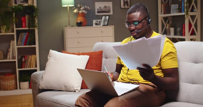 Experienced support specialist cheking documents and consulting client online. Adult Afro American man in headphones working from home during quarantine in evening. Covid-19, epidemic, telework.