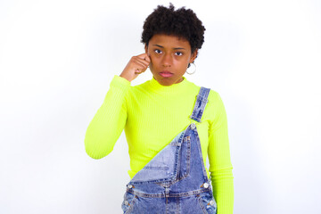 Unhappy young African American woman with short hair wearing denim overall against white wall crying while posing at camera whipping tears with hand.