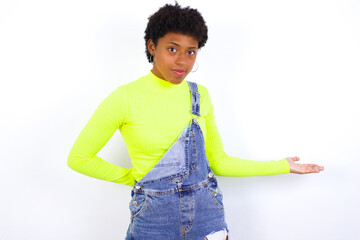 Portrait of young African American woman with short hair wearing denim overall against white wall with arm out in a welcoming gesture.