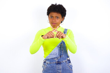 young African American woman with short hair wearing denim overall against white wall has rejection...