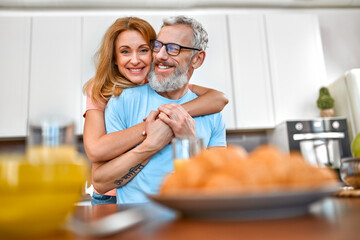 A mature married couple hugs and has breakfast in the kitchen early in the morning and has a good time romantically.
