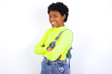 Image of cheerful young African American woman with short hair wearing denim overall against white wall with arms crossed. Looking and smiling at the camera. Confidence concept.