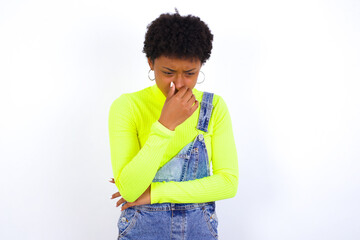 young African American woman with short hair wearing denim overall against white wall, holding his nose because of a bad smell.