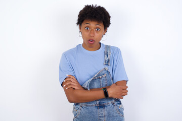 Shocked embarrassed African American female with afro hair wears jeans overalls over white wall keeps mouth widely opened. Hears unbelievable novelty stares in stupor