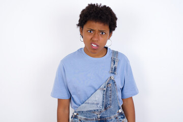 Portrait of dissatisfied African American female with afro hair wears jeans overalls over white smirks face, purses lips and looks with annoyance at camera, discontent hearing something unpleasant