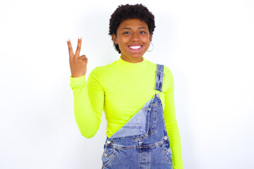 young African American woman with short hair wearing denim overall against white wall smiling and looking friendly, showing number two or second with hand forward, counting down
