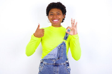 young African American woman with short hair wearing denim overall against white wall smiling and looking happy, carefree and positive, gesturing victory or peace with one hand