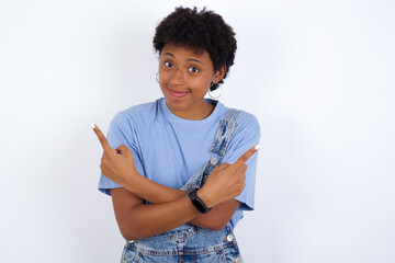 African American female with afro hair wears jeans overalls over white wall crosses arms and points at different sides hesitates between two items or variants. Needs help with decision