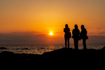 Fototapeta na wymiar 海に沈む夕日を眺める3人の若い女性のシルエット　秋谷海岸　立石公園　梵天の鼻